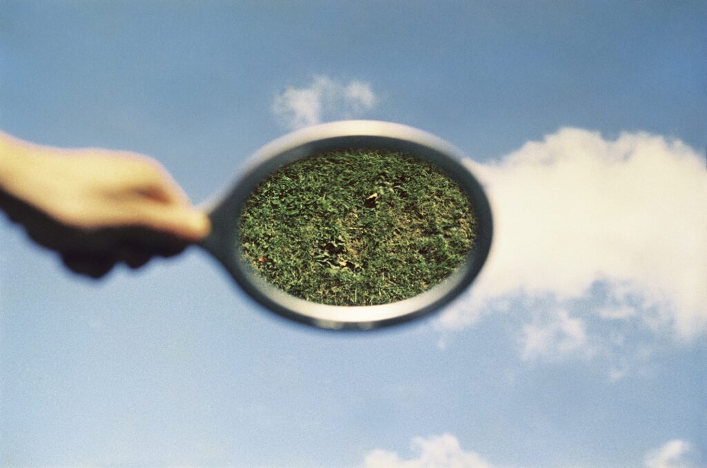 Eine Hand hält einen Spiegel vor einen blauen Himmel mit Wolken, der anstelle des Himmels ein Stück grünes Gras reflektiert