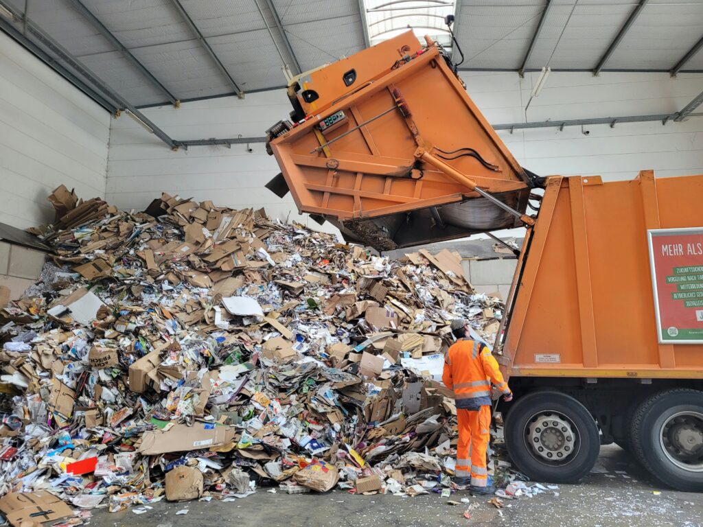 Ein orangefarbener Müllwagen kippt eine große Ladung gemischter Pappe und Papier auf einen riesigen Haufen in einer Recyclinganlage