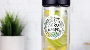 A glass bottle with lemon and lime slices has a "Zero Waste" sticker on it, with an out-of-focus potted plant in the background.