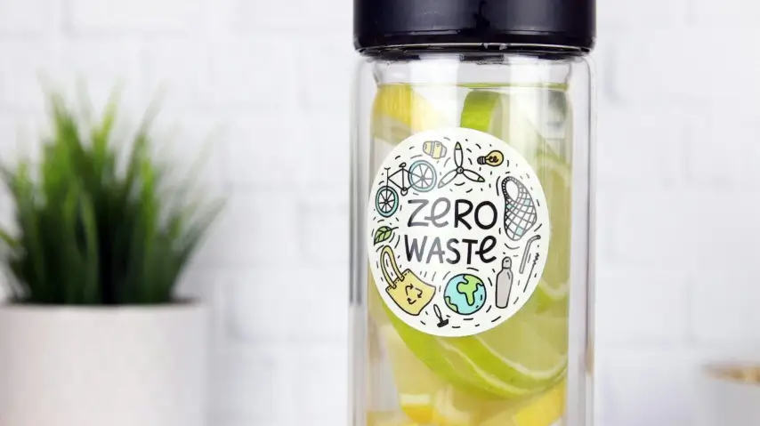 Eine Glasflasche mit Zitronen- und Limettenscheiben hat einen „Zero Waste“-Aufkleber darauf, im Hintergrund eine unscharfe Topfpflanze.